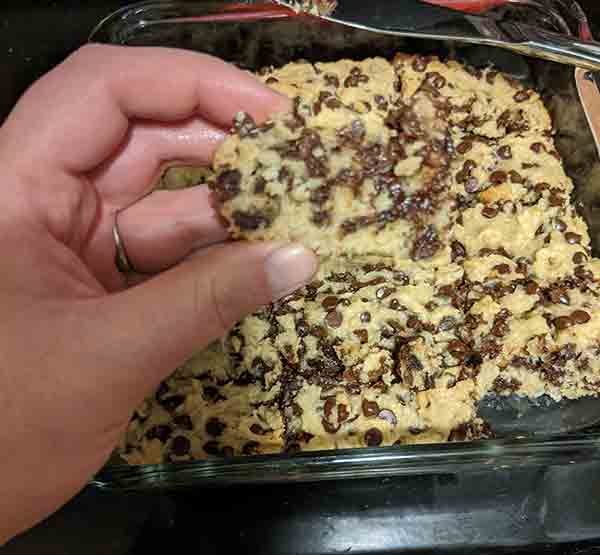 Hand holding up a slice of chocolate chip cookie bar over the pan of bars.