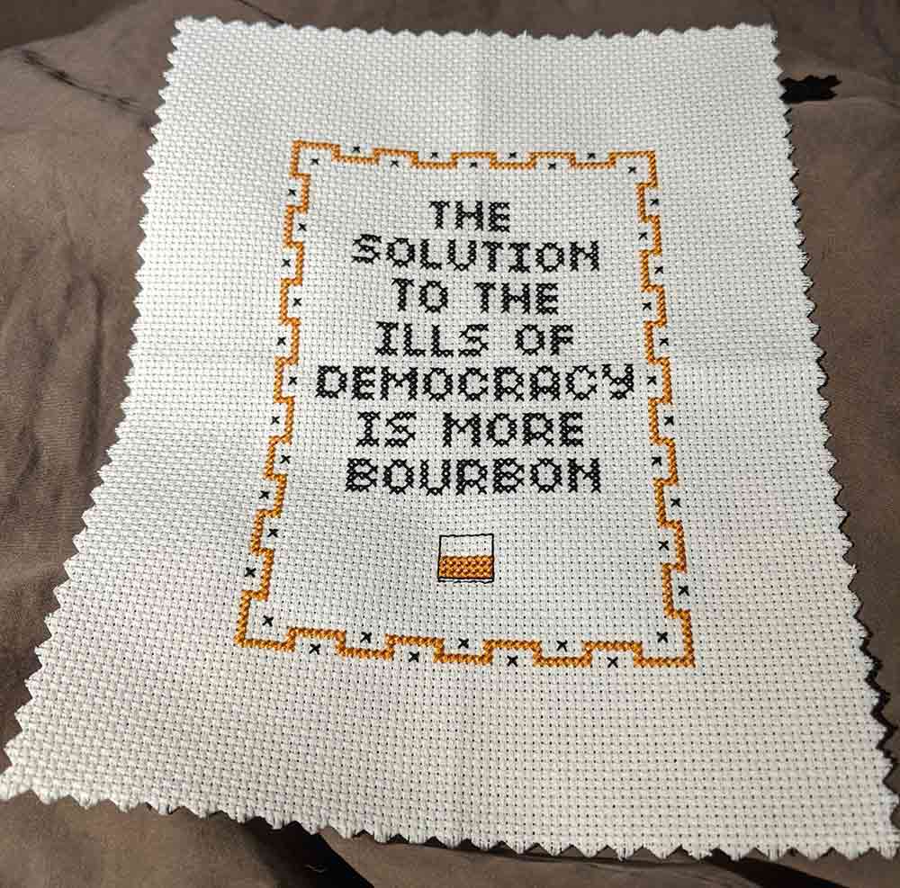 Cross stitch sampler with "The solution to the ills of democracy is more bourbon".