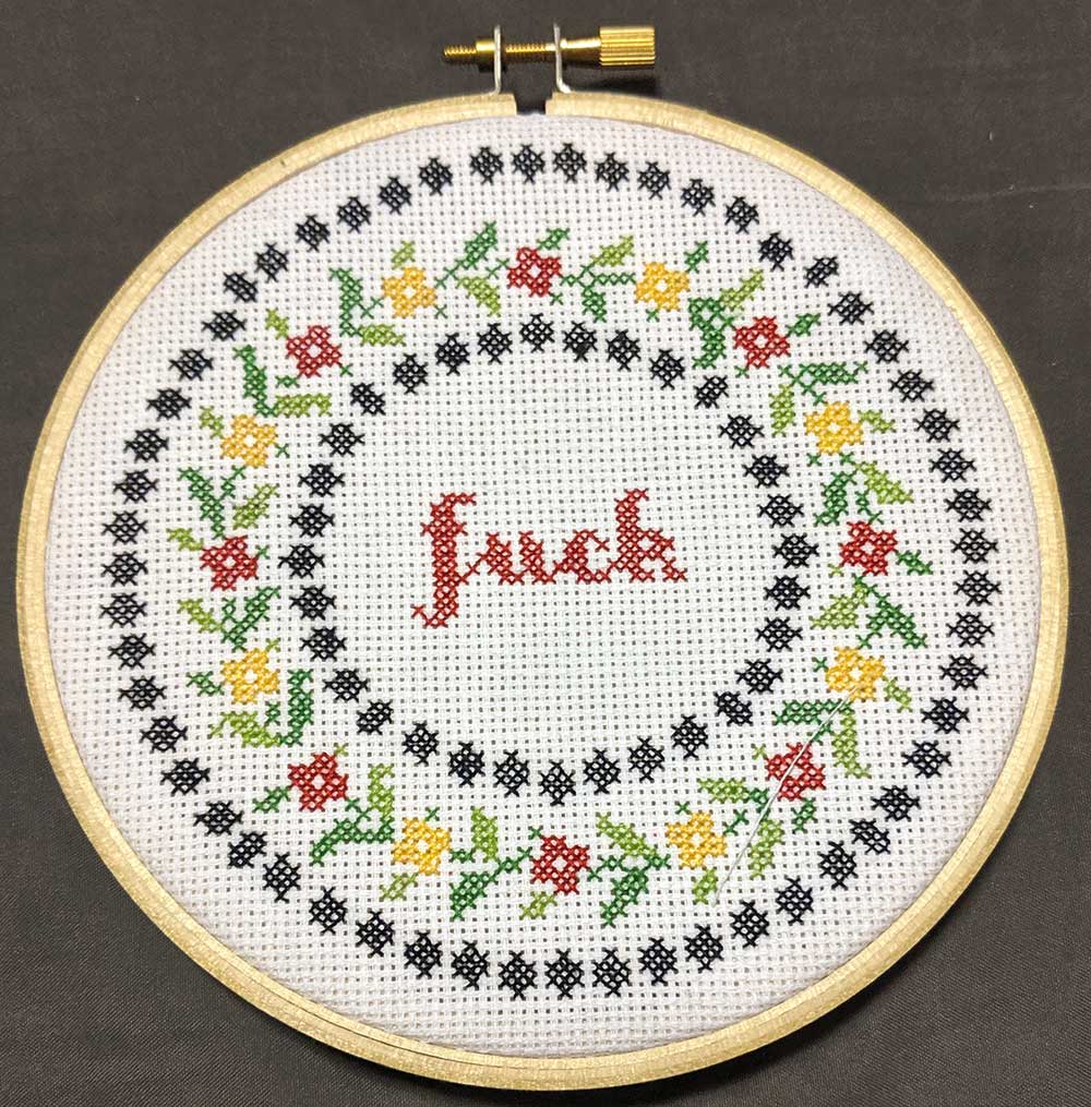 Cross stitch floral wreath with a red cursive "fuck" in the center.