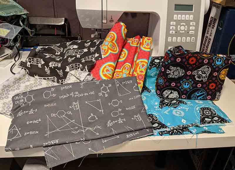 Fabric piled in front of a white sewing machine. Fabric patterns from left to right: grey science equations and diagrams, brown meat cuts, bright geometric, blue Darth Vader, and multicolored Star Wars.