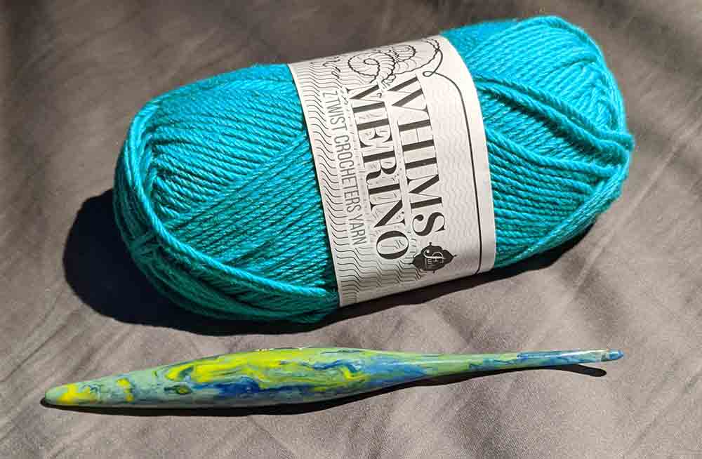 Skein of teal yarn and a green and blue marbled crochet hook on a grey sheet.