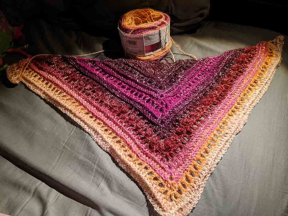 Triangular red, pink, and orange shawl in progress on a grey sheet. The cake of yarn is at the top of the bed above the shawl.